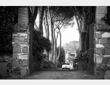 Ingresso di una villa con due pilastri in pietra e cancelli in ferro, che si apre su un vialetto acciottolato fiancheggiato da vegetazione e alti alberi. In fondo si intravede l’edificio della villa e un’auto parcheggiata lungo il viale.