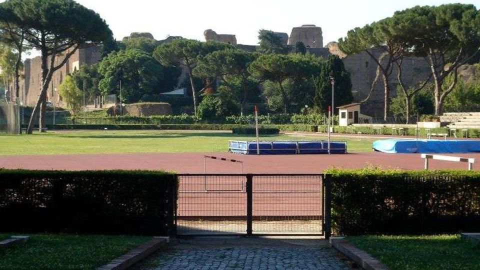 Vista di una pista di atletica all’aperto con pedana per salto in alto e prato interno, delimitata da siepi e un cancello in primo piano. Sullo sfondo si vedono alti pini e mura in pietra di un’area storica.
