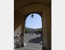 Passaggio ad arco coperto con pareti intonacate e lampione sospeso, che incornicia l’uscita su una piazza pavimentata in pietra. Sullo sfondo si vedono edifici in fila lungo la strada, auto parcheggiate, alberi e colline sotto un cielo sereno.