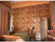 Camera da letto in stile classico con letto in legno e comodini, pareti rivestite da carta da parati a motivi floreali verticali e soffitto decorato con cornici ornate. Sulla sinistra si vede una tenda a righe davanti a una finestra e sulla destra una porta interna.
