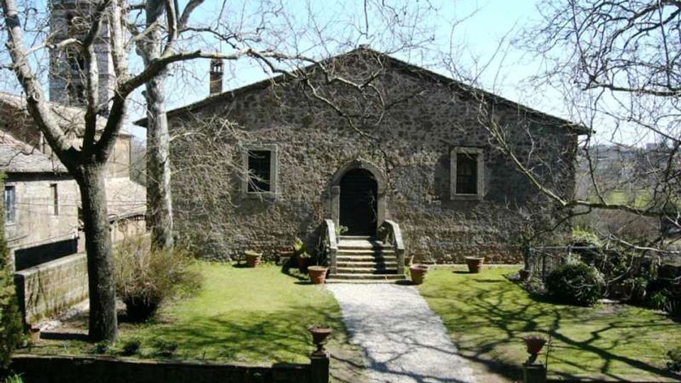 Edificio in pietra a un piano con ingresso ad arco raggiunto da una breve scalinata, affacciato su un giardino con prato e un vialetto centrale. Alberi spogli e vasi in terracotta circondano il percorso, con altri edifici in pietra sullo sfondo.