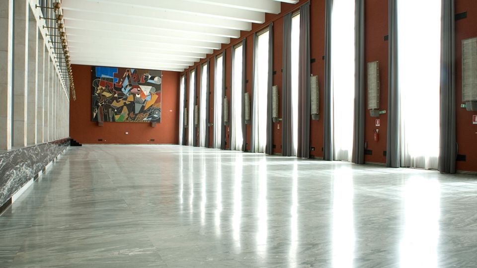 Ampia sala interna con pavimento in marmo lucido e soffitto a travi bianche, affacciata su una lunga fila di alte finestre con tende grigie. Pareti color terracotta con applique e, in fondo, un grande dipinto astratto.