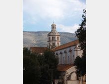 Vista esterna di un complesso monastico in pietra con tetti in tegole rosse e un campanile ottagonale con cupola e croce. L’edificio è circondato da alberi, con colline montuose sullo sfondo sotto un cielo parzialmente nuvoloso.