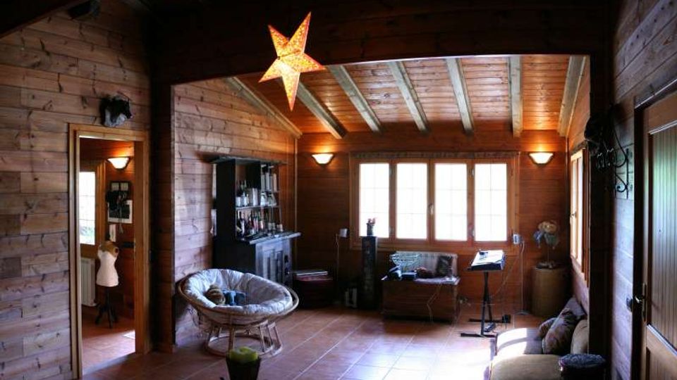 Interno di un soggiorno in legno con soffitto a travi a vista, pavimento in piastrelle e grandi finestre sul fondo. Nella stanza si vedono divano e poltrona, una libreria e una lampada a stella sospesa, con una porta che conduce a un’altra area della casa.