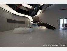 Ampio atrio interno di un edificio moderno, con pavimento chiaro, pareti in cemento e un banco reception bianco curvo al centro. Una grande scala nera a rampe e passerelle sospese attraversa lo spazio sotto un soffitto con pannelli luminosi curvilinei, con ingresso vetrato sul lato destro.