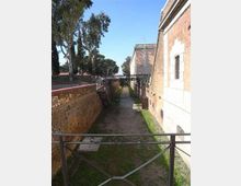 Passaggio pedonale stretto tra alti muri in mattoni e un edificio in muratura, con un fossato erboso e un camminamento centrale. Una ringhiera metallica in primo piano e alberi alti sul lato sinistro incorniciano la vista verso il fondo del corridoio.