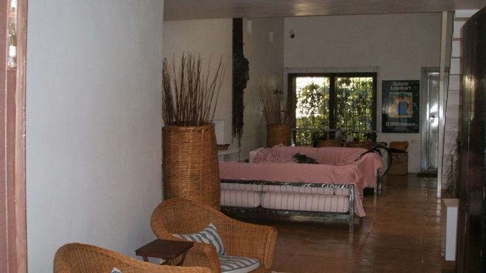 Interno di un’area lounge con pavimento in cotto e pareti chiare, arredata con poltrone in vimini, tavolini e grandi vasi decorativi. Sul fondo si vede un divano coperto in tessuto rosa e una porta a vetri che dà su un’area verde esterna.
