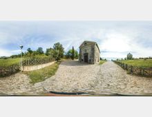 Piazzale in ciottoli accanto a una piccola chiesa in pietra con facciata a timpano e ingresso centrale. Tutt’intorno si vedono campi verdi, alberi e staccionate in legno che delimitano i sentieri sotto un cielo aperto.