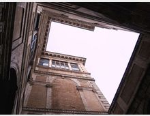 Veduta dal basso di un cortile interno con facciate alte in muratura e pietra, caratterizzate da cornici decorative, finestre rettangolari e un grande riquadro arcuato. Il cielo è visibile attraverso un’apertura superiore irregolare delimitata dai cornicioni del palazzo.