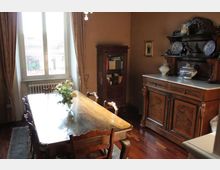 Sala da pranzo con grande tavolo in legno e sedie, illuminata da una finestra alta con tende e radiatore sottostante. Sulla destra c’è una credenza antica con alzata e piatti decorativi, e sul fondo una vetrina in legno con libri.