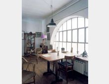 Interno di uno studio con grande finestra ad arco a riquadri che illumina un tavolo da lavoro in legno. Nella stanza si vedono pavimento in assi di legno, sedie in legno e vimini, una lampada a sospensione, una lampada da terra, quadri alle pareti e una vetrina con oggetti.