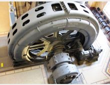 Grande macchinario industriale d’epoca con una massiccia struttura circolare metallica forata e componenti meccanici interni visibili. L’attrezzatura è installata in un ambiente interno con pareti e finiture architettoniche, simile a un museo o a un edificio storico.