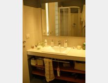 Bagno moderno con lavabo rettangolare su mobile sospeso e grande specchio a parete; sul piano sono disposti vari prodotti da toilette. Sullo sfondo si vede una cabina doccia con porte scorrevoli e pareti piastrellate in tonalità chiare.