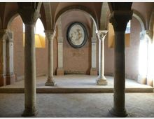 Interno di una sala in pietra con colonne massicce e archi a volta che sostengono il soffitto. Sul fondo, una parete semicircolare ospita un medaglione decorativo, con luce naturale che entra da aperture laterali.