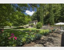 Giardino paesaggistico con prato ampio, aiuole fiorite e vialetto in pietra con tavolini all’aperto, ai piedi di un pendio boscoso. Sullo sfondo si vede una casa in pietra con tetto in coppi, una scalinata e cipressi accanto a un muretto di contenimento.
