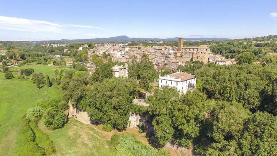 Veduta aerea di un borgo con edifici in pietra e una torre campanaria, circondato da ampie aree verdi e alberate. In primo piano si vede un grande edificio chiaro con tetto in coppi vicino al margine di una scarpata e a campi aperti.