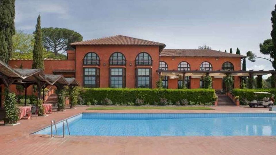 Edificio in mattoni rossi a due piani con tetto in coppi e grandi finestre ad arco, affacciato su un’ampia piscina rettangolare con bordo in cotto. Nell’area esterna ci sono pergolati, siepi curate e alberi alti sullo sfondo.