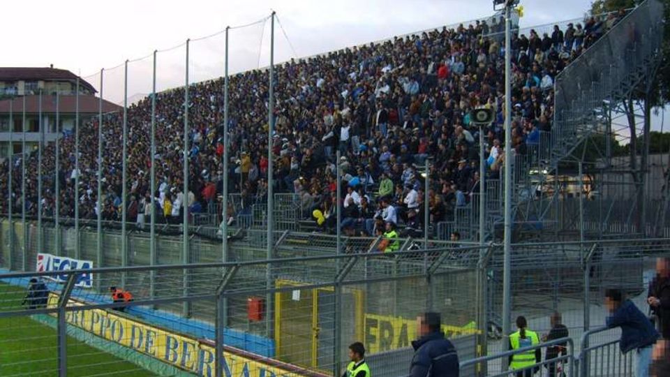 Tribuna gremita di uno stadio di calcio, con gradinate metalliche alte e recinzioni in rete e ringhiere davanti al campo. In primo piano si vedono il bordo del terreno di gioco con cartelloni pubblicitari e strutture di sicurezza lungo la curva.