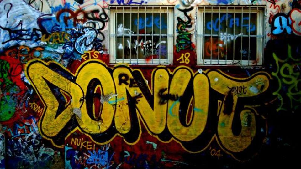 Muro esterno di un edificio ricoperto da graffiti multicolori, con una grande scritta gialla “DONUT” in primo piano. In alto si vedono due finestre rettangolari con inferriate metalliche.