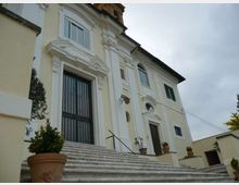 Facciata di un edificio storico in stile barocco, intonacata in giallo chiaro con stucchi bianchi, portale alto con inferriata e finestre ad arco. Davanti, una scalinata in pietra con corrimano e vasi di piante conduce all’ingresso sotto un cielo nuvoloso.
