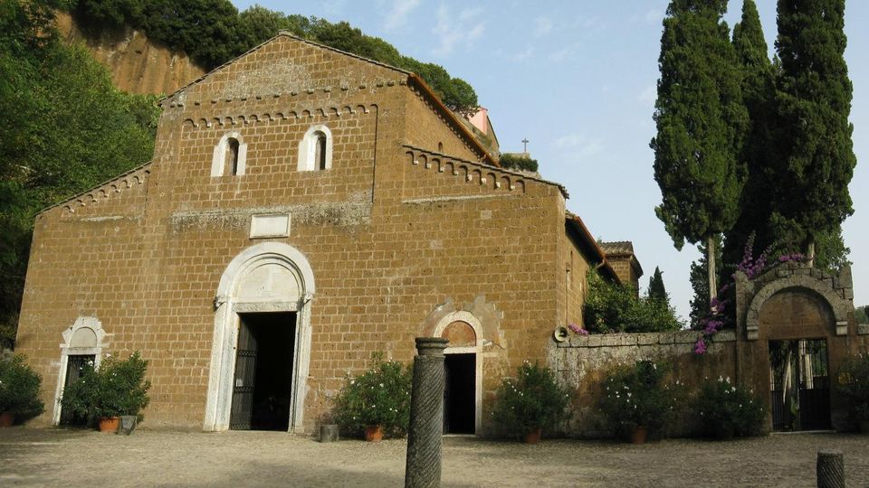 Facciata di una chiesa romanica in pietra con portale ad arco e piccole finestre, affacciata su un ampio piazzale sterrato con vasi di piante. A destra si vede un muro in pietra con ingresso ad arco e alti cipressi, sullo sfondo una collina alberata.