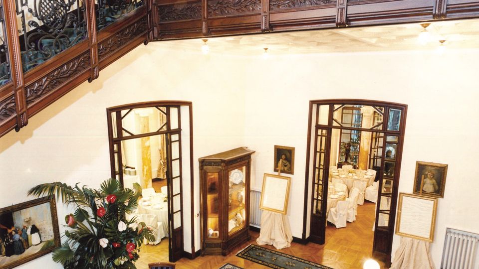Interno di un elegante atrio su due livelli, con balconata in legno e ringhiere in ferro battuto decorato. Al piano terra si vedono pavimento in parquet con tappeti, vetrina espositiva, quadri alle pareti e due porte ad arco che conducono a sale apparecchiate per banchetti.