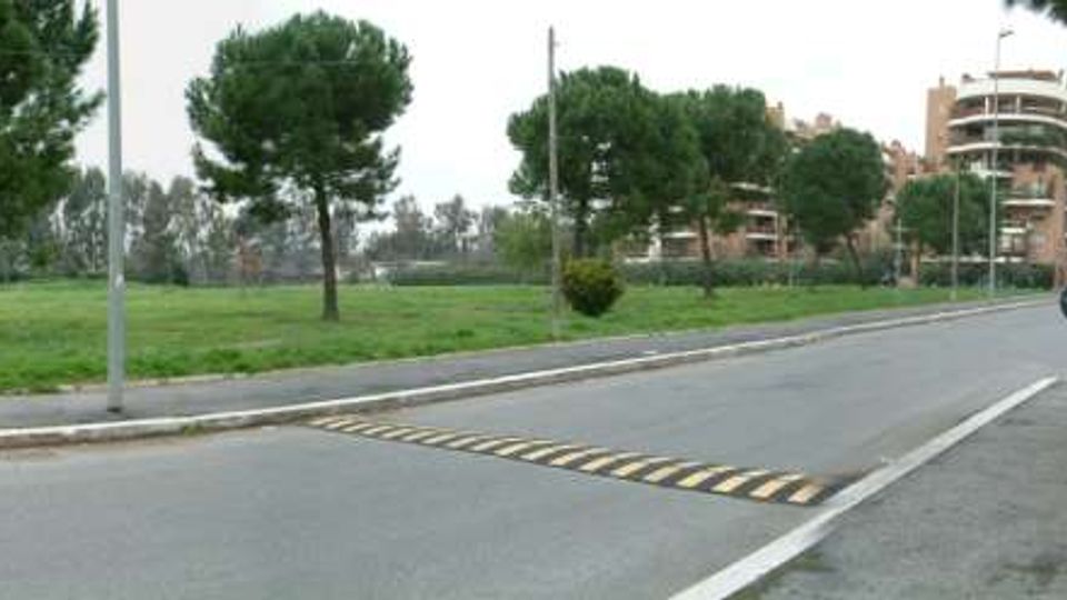 Strada urbana con rotatoria e attraversamento rialzato, affiancata da un’ampia area verde con pini. Sullo sfondo si vedono edifici residenziali multipiano e lampioni lungo la carreggiata.