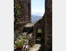 Stretto vicolo in pietra tra edifici rustici, con muri in pietra a vista e piccole piante lungo il percorso. In fondo si vede un passaggio ad arco con cancello e, oltre, una vista sulle montagne sotto un cielo sereno.