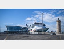 Terminal portuale moderno con facciata vetrata e tetto curvo, accanto a una grande nave da crociera ormeggiata. Sul lato destro si vede una torre in pietra tipo faro e, in primo piano, un ampio piazzale asfaltato con segnaletica.