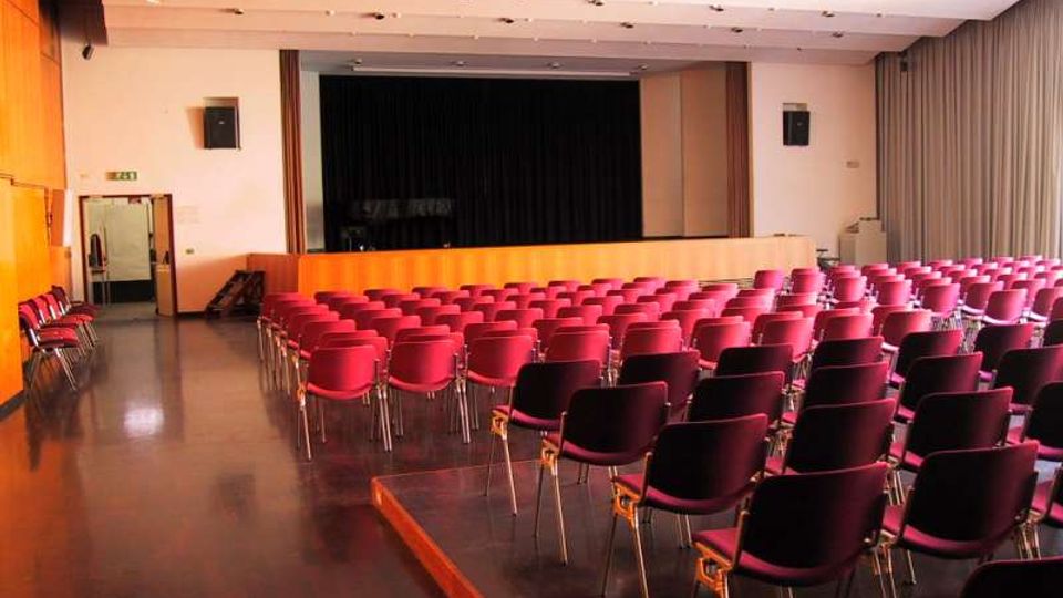 Sala auditorium con file di sedie rosse e nere rivolte verso un palco rialzato con sipario scuro. Pareti chiare con pannelli in legno, illuminazione a soffitto e uscita laterale sulla sinistra.