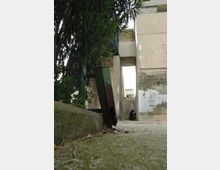 Vicolo stretto tra edifici in cemento con muri scrostati e macchie di umidità, visto dal livello del suolo. In primo piano si vedono vegetazione e un contenitore metallico arrugginito, con pavimentazione in cemento e un’apertura luminosa in fondo.