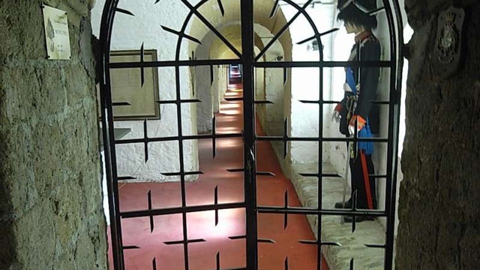 Ingresso ad arco in mattoni con cancello in ferro a grata, che conduce a un lungo corridoio sotterraneo illuminato, con pavimento rosso e muri bianchi. Lungo il passaggio si susseguono archi e nicchie, con pannelli informativi alle pareti.