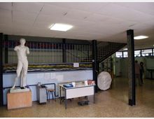 Interno di un laboratorio scolastico o artistico con pavimento in graniglia, soffitto a pannelli e luci al neon, con scrivanie e attrezzature. A sinistra c’è una statua classica in gesso su basamento, mentre sullo sfondo si vedono una scala metallica e una ringhiera che affacciano su un soppalco.