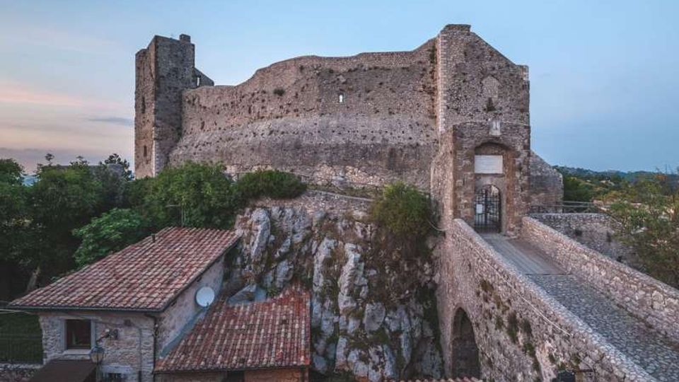 Castello in pietra su uno sperone roccioso, con mura alte e torri rettangolari, collegato da un ponte in muratura che conduce a un portale d’ingresso ad arco. In primo piano si vedono tetti in coppi di edifici sottostanti e vegetazione circostante, con cielo sereno sullo sfondo.