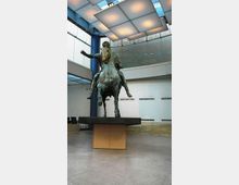 Atrio interno di un museo moderno con grandi superfici vetrate e pannelli luminosi sospesi sul soffitto. Al centro, su un basamento, una grande statua equestre in bronzo circondata da pareti chiare e una breve scalinata sullo sfondo.