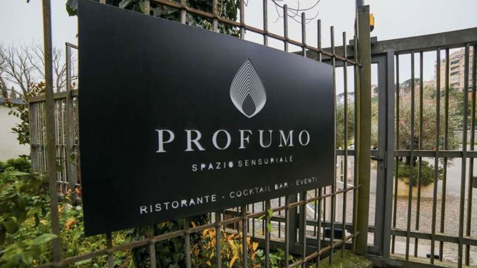 Insegna nera con il logo e la scritta “PROFUMO – Spazio sensoriale” fissata a una recinzione metallica vicino a un cancello d’ingresso. Sullo sfondo si vedono vegetazione, un vialetto e edifici lontani.