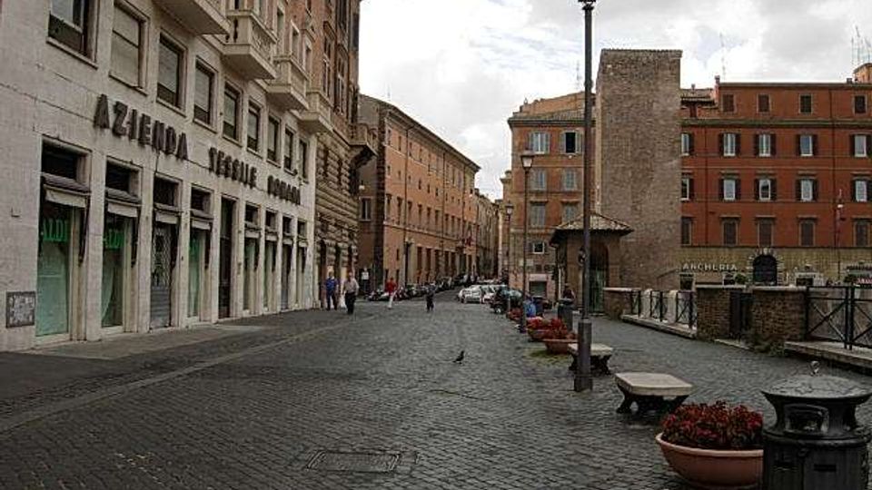 Strada urbana lastricata con palazzi in muratura su entrambi i lati, lampione centrale e marciapiede con panchine e fioriere. A sinistra si vede un edificio con grandi vetrine e l’insegna “Azienda Elettrica Romana”, mentre sulla destra c’è una ringhiera lungo un’area rialzata con altri edifici sullo sfondo.