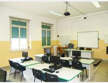 Aula informatica con banchi disposti in file, postazioni con monitor e sedie nere, pareti bianche e gialle e pavimento grigio. Sul fondo ci sono una cattedra, una lavagna bianca e un proiettore a soffitto, con finestre laterali dotate di tapparelle verdi.