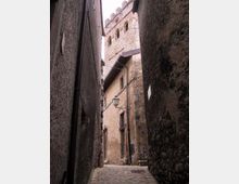 Vicolo stretto in pietra tra edifici alti con pareti in muratura grezza e selciato irregolare. In fondo si vede una torre in pietra con merli e finestre ad arco, con un lampione a parete e cavi sospesi lungo la facciata.