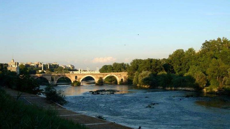 Veduta di un fiume con un ponte in pietra ad arcate al centro, circondato da alberi e vegetazione sulle rive. Sullo sfondo si intravedono edifici della città sotto un cielo sereno.