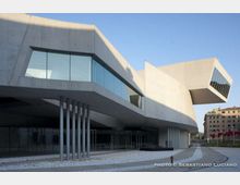 Edificio moderno in cemento e vetro con volumi sporgenti e grandi finestre angolari, sostenuto da sottili pilastri metallici. Davanti si vede una piazza pavimentata con percorso curvo, mentre sullo sfondo compaiono altri palazzi urbani.