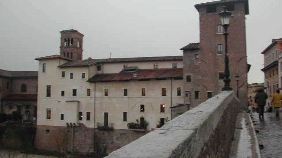 Vista da un ponte in pietra con parapetto e lampione su un complesso di edifici storici lungo un fiume, con facciate intonacate chiare e una torre in mattoni. Sullo sfondo si vede un campanile e i tetti in coppi sotto un cielo grigio.