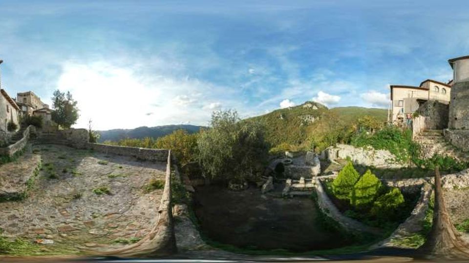 Panorama di un piccolo borgo in pietra con terrazze e muri in muratura che scendono verso un’area centrale incassata. Sullo sfondo si vedono colline boscose e montagne sotto un cielo azzurro con nuvole sparse.