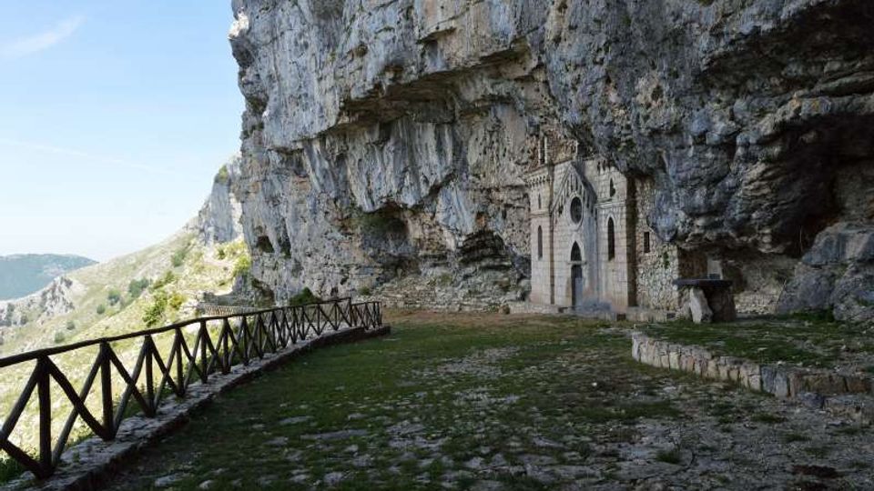 Piccola chiesa in pietra con facciata gotica incastonata sotto una grande parete rocciosa a strapiombo, accanto a una terrazza erbosa e pavimentata. Un camminamento con staccionata in legno segue il bordo della scogliera con vista sulle montagne circostanti.