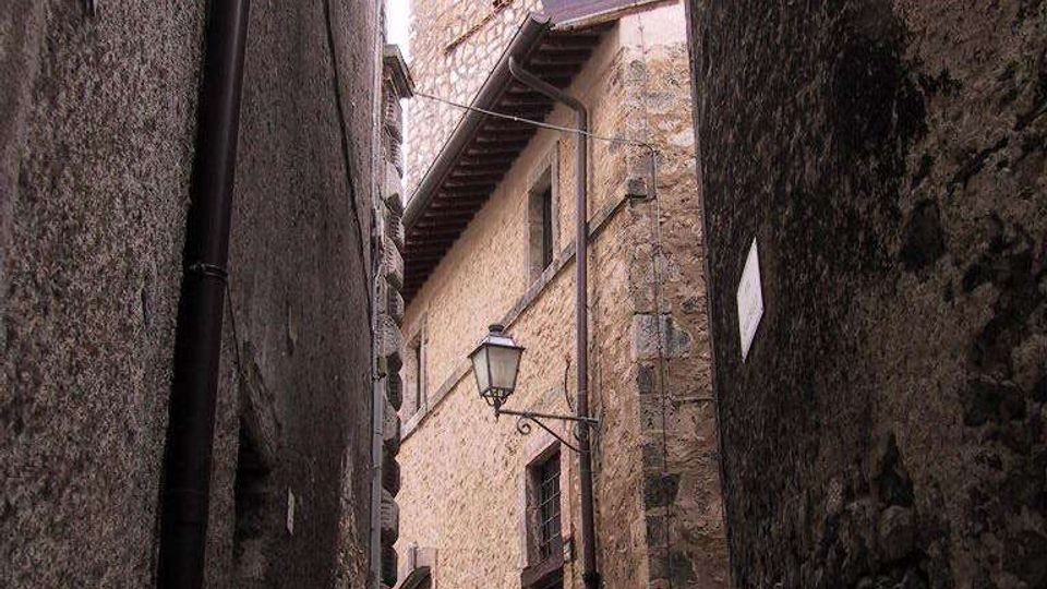 Vicolo stretto in pietra tra edifici alti con pareti in muratura grezza e selciato irregolare. In fondo si vede una torre in pietra con merli e finestre ad arco, con un lampione a parete e cavi sospesi lungo la facciata.