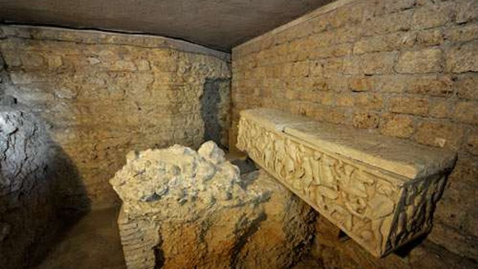 Interno sotterraneo in pietra con soffitto basso e pareti in blocchi, illuminato debolmente. Sulla destra c’è un sarcofago in pietra con rilievi decorativi e al centro un basamento con frammenti di pietra.