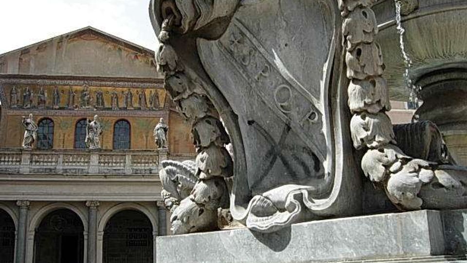 Dettaglio ravvicinato di una grande fontana in pietra scolpita, con elementi decorativi curvilinei e una vasca circolare sul lato destro. Sullo sfondo si vede un edificio storico con portico ad archi, statue e facciata con mosaici.