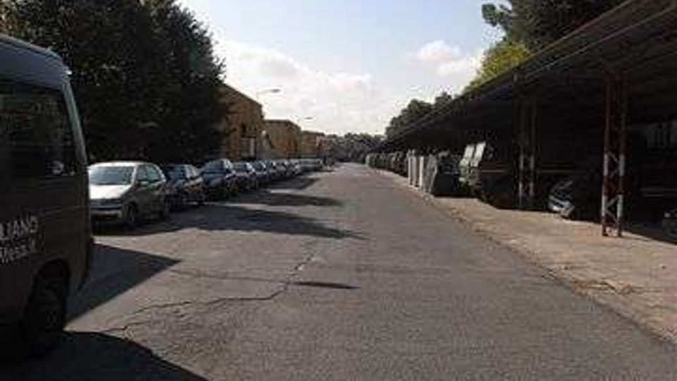 Strada urbana asfaltata e dritta con auto parcheggiate lungo entrambi i lati. A destra si vede una lunga tettoia con struttura metallica e alberi alti sullo sfondo, sotto un cielo sereno.