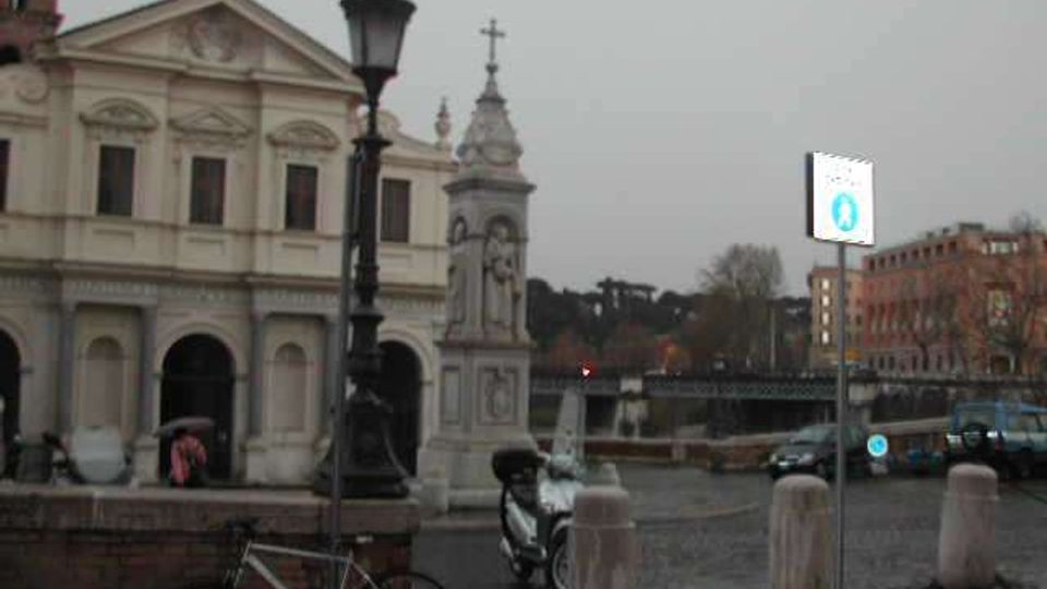 Piazza urbana con una chiesa storica dalla facciata classica e un’edicola monumentale con croce; in primo piano si vedono lampione, bicicletta e dissuasori. Sullo sfondo compaiono una strada con auto, un ponte e palazzi lungo il fiume.