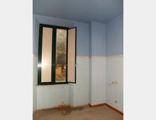 Interno di una piccola stanza vuota con pareti bicolore azzurro e fascia decorativa, pavimento piastrellato e zoccolino arancione. Finestra a due ante con telaio verde e una parte aperta, affacciata sull’esterno.